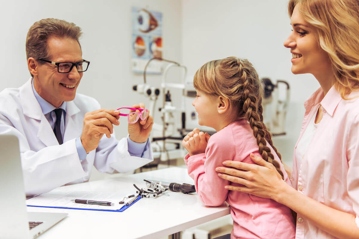 Male pediatric optometrist showing pink glasses to a young girl and her mother